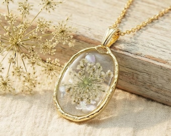 May include: A gold-colored necklace with an oval pendant. The pendant contains a dried flower encased in clear resin. The necklace chain is also gold. A dried flower head is next to the necklace on a wooden surface.