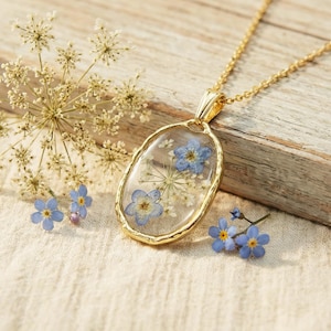 May include: A gold pendant necklace featuring an oval resin pendant with blue and white dried flowers. The necklace is displayed on a wooden surface with additional flowers. The chain is gold.