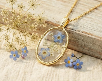 May include: A gold-toned oval pendant necklace featuring pressed blue forget-me-not flowers and white lace-like botanical elements encased in a clear resin frame. The pendant hangs from a delicate gold-toned chain, showcasing a botanical design.