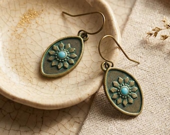 May include: A pair of bronze-toned oval drop earrings featuring a raised floral pattern with a small, round, light blue stone at the center of each flower. The earrings have curved metal ear wires for attachment and a textured finish.