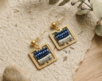 May include: A pair of gold-toned square earrings featuring a horizontal row of small beads in shades of dark blue, light blue, white, and grey. The earrings have a post-back closure and a textured metal frame surrounding the beaded center section.