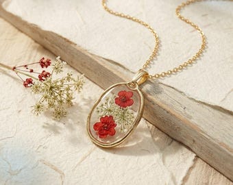 May include: A gold-toned oval pendant necklace featuring pressed red flowers and white lace-like botanical elements encased in clear resin. The pendant hangs from a delicate gold-toned chain, accompanied by a small sprig of matching dried flowers.
