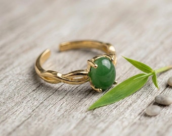 May include: A gold-toned ring featuring a smooth, oval-shaped green stone set in a prong setting. The band has a twisted, open design that allows for adjustment. The ring is positioned next to a single green leaf and small gray pebbles.