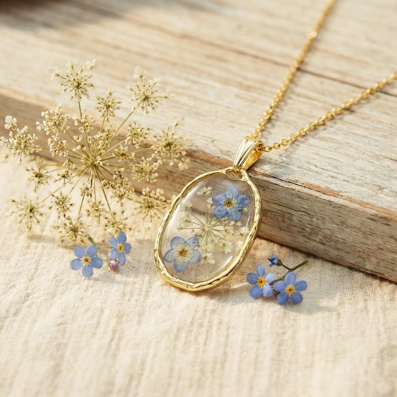 May include: A gold-colored pendant necklace with an oval-shaped, clear resin pendant. The pendant contains blue and white dried flowers. The necklace is displayed on a wooden surface with additional flowers.