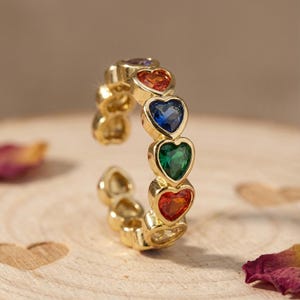 May include: A gold-colored ring featuring a series of heart-shaped gemstones in a rainbow of colors. The gemstones include red, orange, blue, and green. The ring is set on a wooden surface with a blurred background.