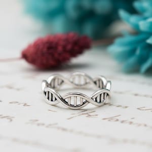 May include: A silver-toned ring featuring a continuous, repeating DNA double helix structure around the entire band. The metal has a polished finish with darkened recessed areas between the vertical bars to highlight the intricate molecular design.