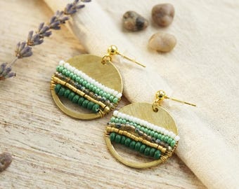 May include: A pair of gold-colored, round earrings. Each earring features a beaded design with rows of white, green, and gold beads. The earrings have a post-back closure. The background includes a sprig of lavender and small stones.