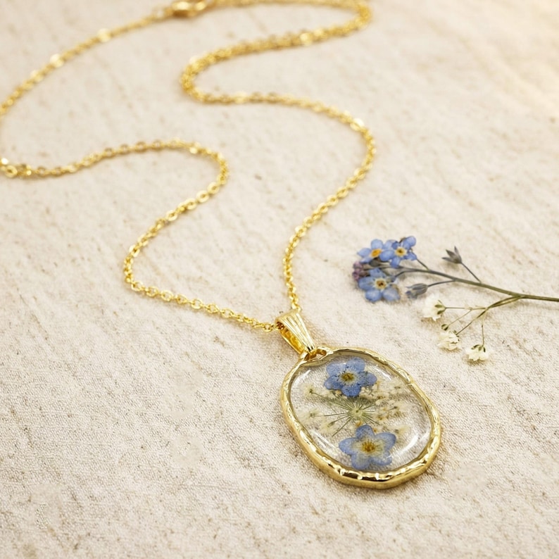 May include: Gold-toned necklace with an oval pendant. The pendant features blue and white dried flowers encased in clear resin. The chain and pendant frame have a textured finish. A sprig of blue flowers rests beside the necklace.