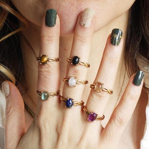 May include: A hand model wearing gold rings with different colored gemstones. The rings are all set in a twisted band design. The gemstones include tiger's eye, black onyx, white moonstone, carnelian, aquamarine, lapis lazuli, and amethyst.