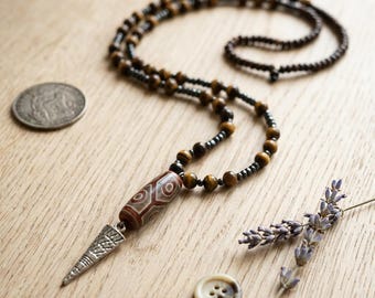May include: A beaded necklace featuring alternating brown tiger eye beads and small dark metallic beads. The necklace includes a cylindrical patterned bead and a silver-toned triangular pendant with etched geometric lines and details.