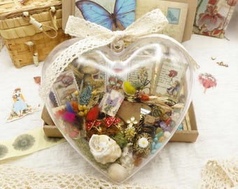 May include: A clear, heart-shaped container filled with various decorative elements, including dried flowers, seashells, and vintage ephemera. A large blue butterfly and a cream-colored lace bow adorn the top. The container is filled with colorful items.