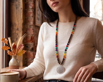 May include: A long necklace featuring dark brown wooden beads interspersed with multi-colored gemstone beads in a repeating pattern. The necklace includes a large, circular silver-toned pendant with an intricate geometric flower of life pattern.