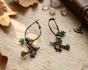 May include: A pair of hoop earrings in an antique bronze finish, each featuring a dangling charm of a witch riding a broomstick. The hoops are accented with small, round green and dark red beads, creating a detailed accessory with a metallic tone.