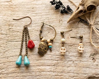 May include: A collection of four earrings featuring bronze-toned metal chains and hoops. The set includes a drop earring with two turquoise-colored teardrop beads and a red bead, a hoop earring with a skull charm, and two stud earrings with small skull beads.