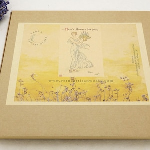 May include: A brown cardboard box with a decorative label featuring an illustration of a woman holding flowers. The label has the text "Here's flowers for you" and the website address "www.verdaartisanworks.com". A bundle of dried lavender is on the left.