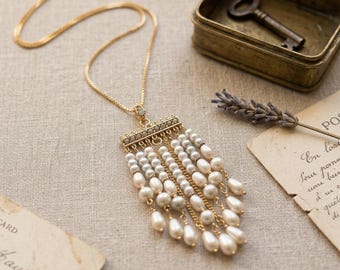 May include: A gold-tone necklace featuring a rectangular pendant with multiple dangling strands of white and cream-colored teardrop-shaped pearls. The pendant top is embellished with small clear stones, and the chain is a delicate gold-tone metal link.