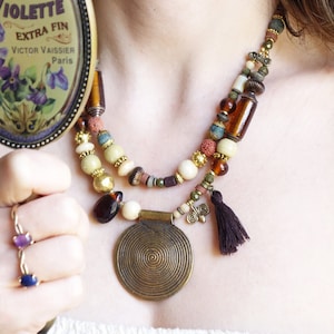 May include: A layered necklace with a large brass pendant, featuring a spiral design. The necklace is made of various beads, including amber, white, brown, and green. A small tassel hangs from the pendant.