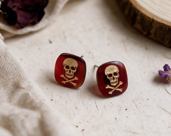 May include: A pair of square stud earrings featuring a deep red translucent base with a gold-toned skull and crossbones emblem centered on each piece. The earrings have a smooth, rounded edge design and include metal posts for secure attachment.