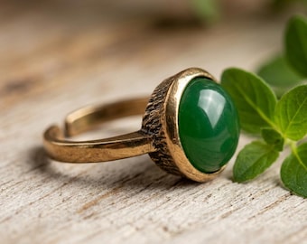 May include: A bronze-toned metal ring featuring a smooth, round, dark green cabochon gemstone set within a textured, rugged bezel. The band is open at the back, showing a metallic finish with visible signs of wear and a dark, aged patina.