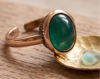 May include: An open-style ring featuring a polished, oval-shaped dark green gemstone set in a hammered copper band. The metal surface displays a textured, rustic finish around the bezel, contrasting with the smooth, rounded green stone.