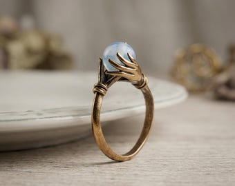 May include: A bronze-toned metal ring featuring a band that splits into two stylized hands with elongated fingers. The hands are positioned to cradle a single, smooth, pale blue spherical stone at the center of the ring setting.