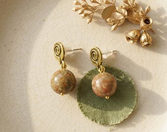 May include: A pair of earrings featuring a gold-toned metal spiral stud attached to a round, speckled, earth-toned stone bead. The earrings are displayed alongside a gold-toned metal botanical sprig and a small, circular, olive green fabric patch.