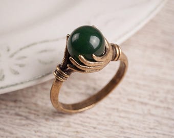 May include: A ring featuring a polished dark green spherical stone held by a metallic bronze-toned setting shaped like a hand. The band has a textured, antique finish with ribbed detailing on the sides, creating a distinct and detailed appearance.