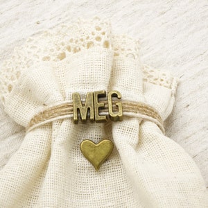 May include: A small, off-white fabric bag with a lace trim at the top. The bag is tied with twine and features the name "MEG" in bronze letters. A bronze heart charm is attached below the name.