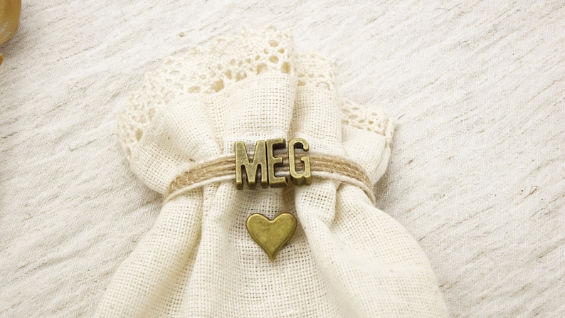 May include: A small, off-white fabric bag with a lace trim at the top. The bag is tied with twine and features the name "MEG" in bronze letters. A bronze heart charm is attached below the name.