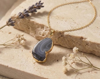 May include: A gold chain necklace featuring a teardrop-shaped pendant with a polished grey agate stone. The stone displays natural concentric banding in shades of dark and light grey, encased in a thin gold-toned metal rim with a small bail.