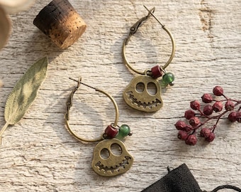 May include: A pair of bronze-toned hoop earrings featuring a dangling skull charm with a wide, toothy grin. Each earring is accented with one small red bead and one small green bead attached to the hoop, showcasing a metallic finish.