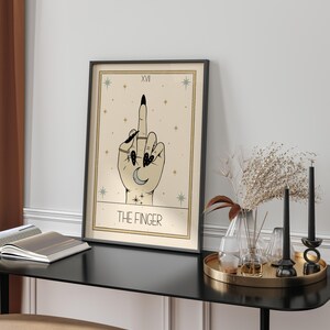 Puede incluir: Una carta del tarot con fondo beige y estrellas negras. La carta representa una mano con esmalte de u&ntilde;as negro haciendo el dedo medio. La carta se titula "The Finger" y tiene el n&uacute;mero XVI en la esquina superior izquierda.