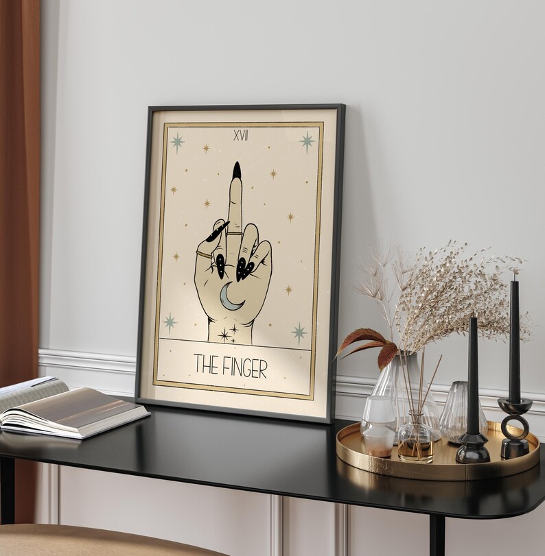 Puede incluir: Una carta del tarot con fondo beige y estrellas negras. La carta representa una mano con esmalte de u&ntilde;as negro haciendo el dedo medio. La carta se titula "The Finger" y tiene el n&uacute;mero XVI en la esquina superior izquierda.