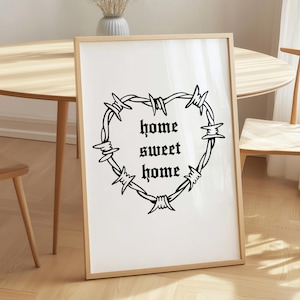 May include: A framed print with a heart-shaped barbed wire design and the words "home sweet home" in a gothic font. The print is displayed on a wooden floor. The frame is light-colored.