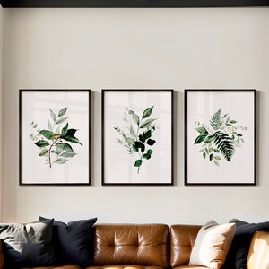 May include: Three framed watercolour prints of green foliage, each with a different arrangement of leaves and branches. The prints are displayed on a white wall above a brown leather sofa.