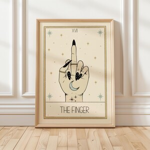 Puede incluir: Una carta del tarot con fondo beige y estrellas doradas. La carta se titula "The Finger" y presenta una mano con esmalte de u&ntilde;as negro haciendo el dedo medio. La mano tiene una luna creciente en la palma.