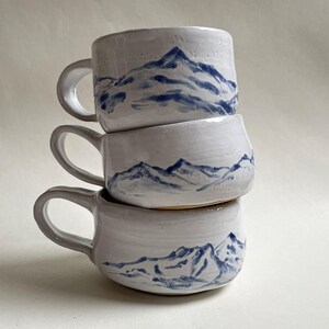 Könnte beinhalten: Drei weiße Keramikbecher, die übereinander gestapelt sind. Jeder Becher hat ein handgemaltes blaues Bergketten-Design. Die Becher haben abgerundete Körper und gebogene Griffe, ideal für Heißgetränke.
