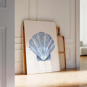 May include: A framed print with a blue and white seashell design. The artwork is propped against a white wall, with a gold frame. The image is in a room with wooden flooring and an open doorway, with a glimpse of a sofa.