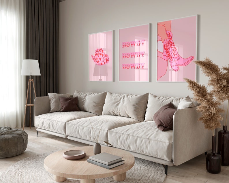 Op de afbeelding: Drie roze ingelijste prints met witte passe-partouts die aan een witte muur hangen boven een lichtbeige bank. De prints tonen een cowboyhoed, het woord "Howdy" drie keer herhaald en een paar roze cowboylaarzen.