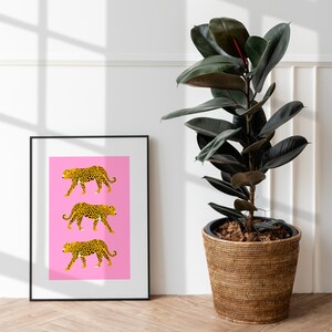 Op de afbeelding: Een ingelijst print met een roze achtergrond en drie zwarte en gele luipaard silhouetten die in een rij lopen. De print hangt aan een witte muur naast een kamerplant in een geweven mand.