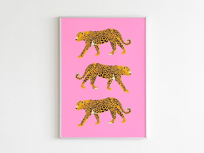 Op de afbeelding: Drie gestileerde luipaarden in een wandelende pose zijn afgedrukt op een roze achtergrond. De luipaarden zijn geel met zwarte vlekken. De afbeelding is ingelijst in een zilveren lijst.