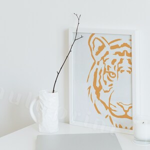 Puede incluir: Un marco blanco con una impresi&oacute;n minimalista de la cara de un tigre en un color naranja apagado. La impresi&oacute;n est&aacute; sobre un fondo blanco. Un jarr&oacute;n blanco con una superficie texturizada contiene una rama sin hojas. Una vela blanca est&aacute; sobre una superficie blanca.