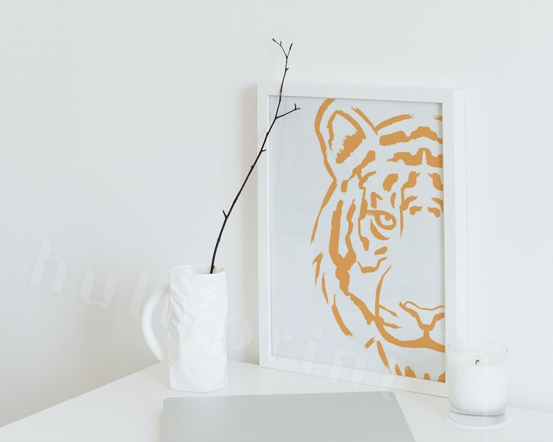 Puede incluir: Un marco blanco con una impresi&oacute;n minimalista de la cara de un tigre en un color naranja apagado. La impresi&oacute;n est&aacute; sobre un fondo blanco. Un jarr&oacute;n blanco con una superficie texturizada contiene una rama sin hojas. Una vela blanca est&aacute; sobre una superficie blanca.