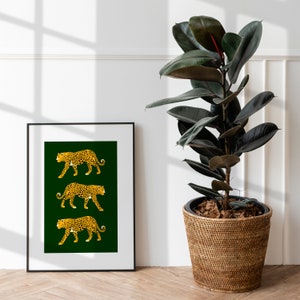 May include: A framed print with a green background and three gold leopard silhouettes. The print is hanging on a white wall next to a potted rubber plant.