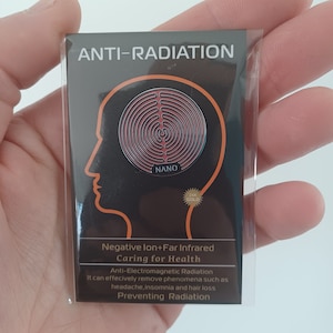 Peut inclure: Un autocollant noir et argent avec le texte "ANTI-RADIATION" et "NANO" en blanc. L'autocollant présente un motif circulaire rouge et argent avec une étoile dorée et le texte "24K GOLD". Le texte "Negative Ion+Far Infrared Caring for Health Anti-Electromagnetic Radiation It can effectively remove phenomena such as headache, insomnia and hair loss Preventing Radiation" est imprimé en blanc sur fond noir.