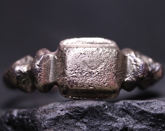 May include: A silver-toned metal ring featuring a central square-shaped face with a textured, matte surface. The band displays a series of raised, geometric decorative elements extending from the sides of the square setting toward the shank.