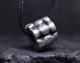 May include: A silver-toned cylindrical pendant with a textured, porous surface featuring raised circular bumps, suspended on a thin black braided cord. The metal piece has a matte, weathered finish with visible indentations and a rough, uneven exterior.