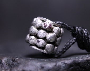 May include: A cylindrical silver-toned bead with a textured surface composed of multiple small, raised circular bumps. The bead features small dark reddish-brown inclusions between the bumps and is threaded onto a black braided cord.