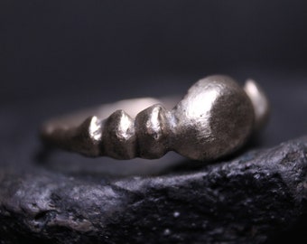 May include: A silver-toned metal ring featuring a textured, uneven band with a series of rounded, raised protrusions along the shank and a larger, smooth, oval-shaped focal point at the top, displaying a matte, weathered finish throughout.