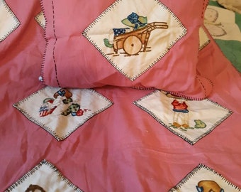 May include: A pink quilted blanket with embroidered images of children and a wagon. The blanket is made of cotton and has a soft, comfortable feel.
